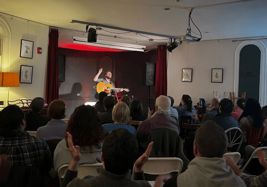 Michael Maloney plays guitar on stage in front of a crowd of clapping audience members.